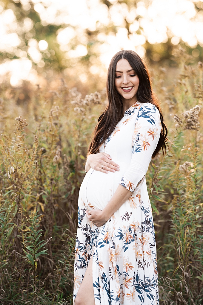 pregnant woman in white floral dress stands in green field at sunset