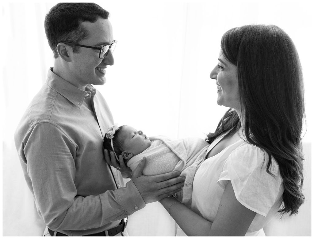 mom and dad hold newborn girl in front of studio window in black and white