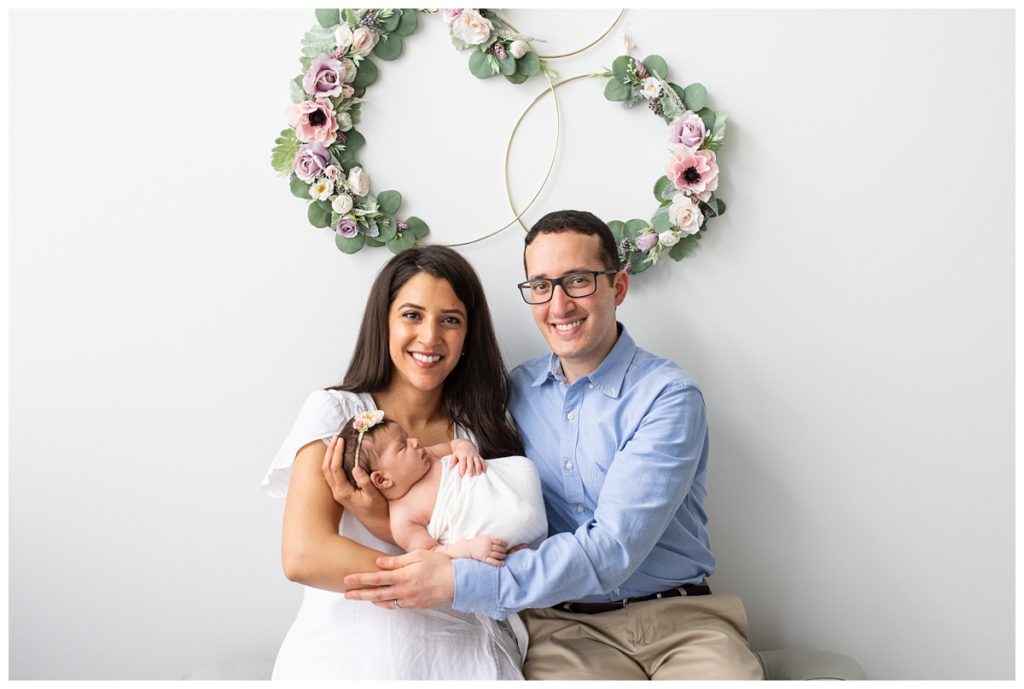 mom and dad hold newborn girl behind wreath of flowers in studio