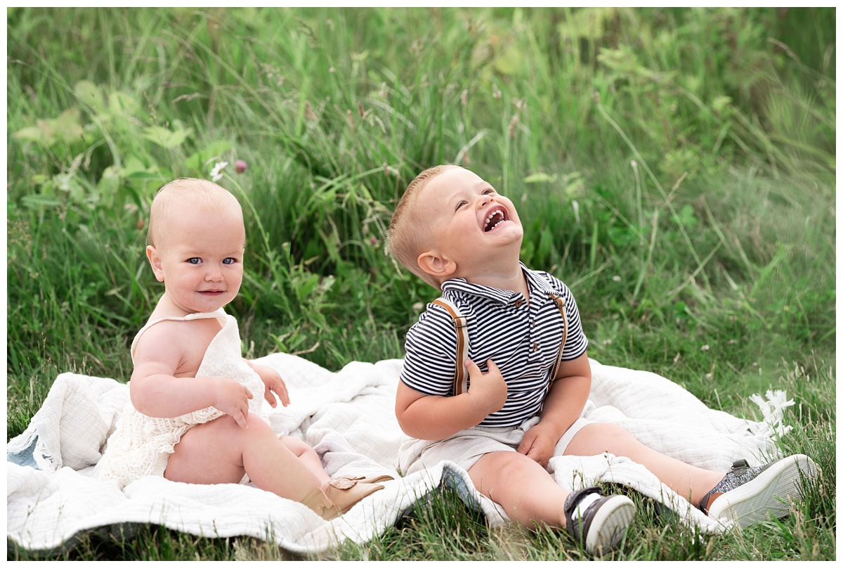 toddler brother and baby sister sit in field and laugh