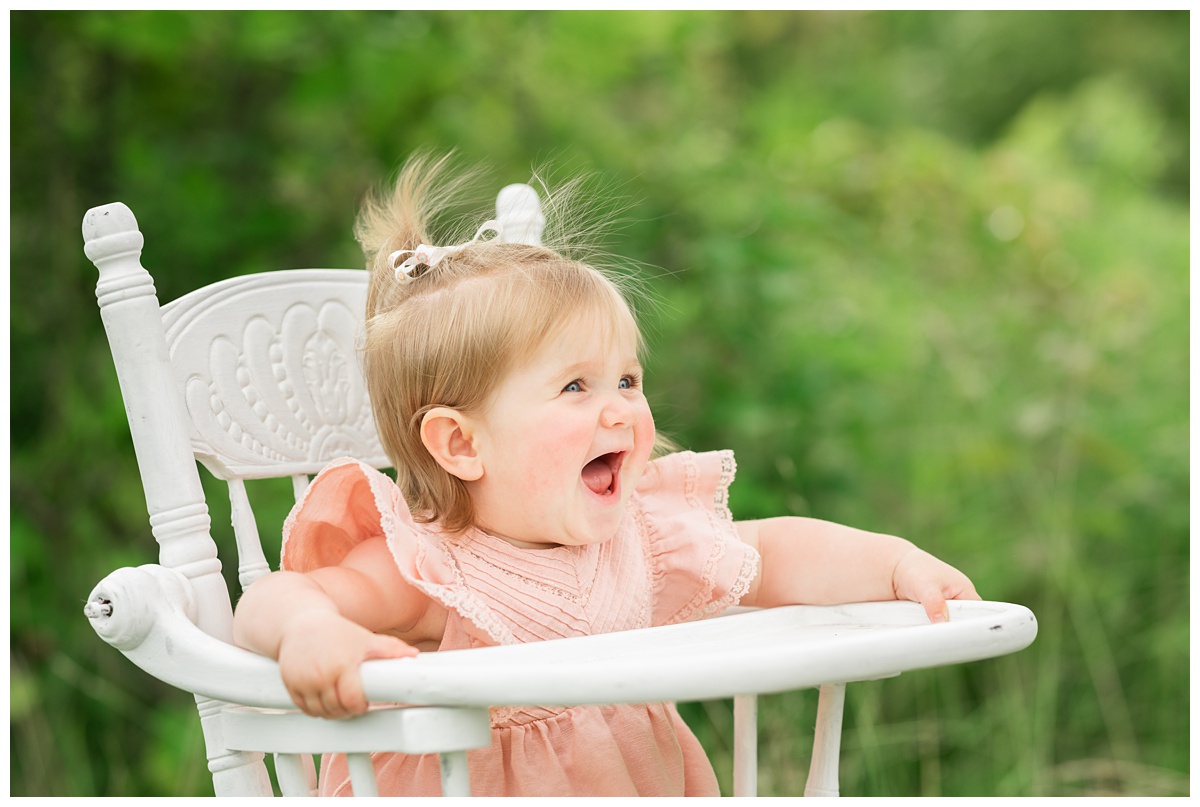 Family Photographers Columbus baby girl holds highchair and smiles big in green field