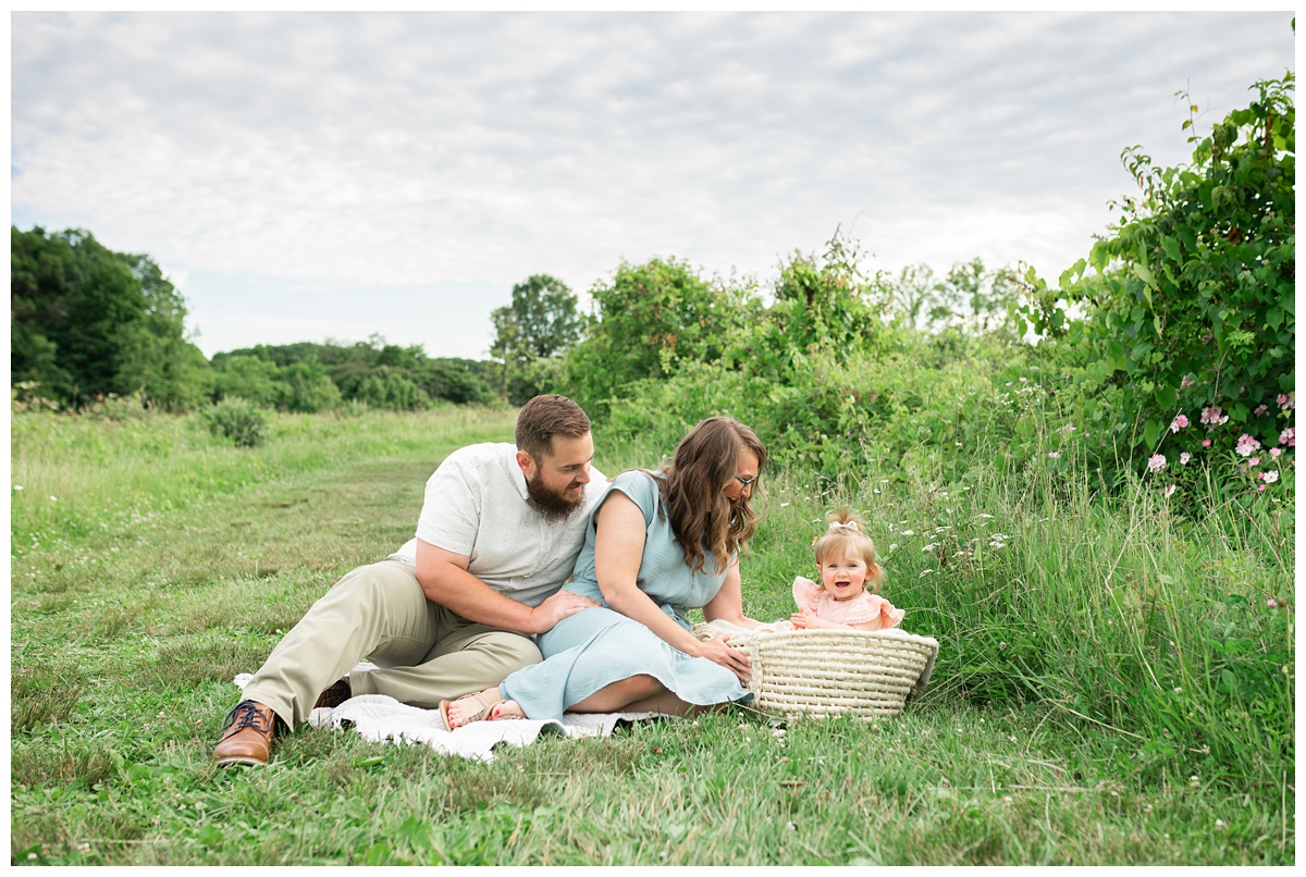 Family Photographers Columbus family of three sit in green field with wild flowers and blue cloudy sky