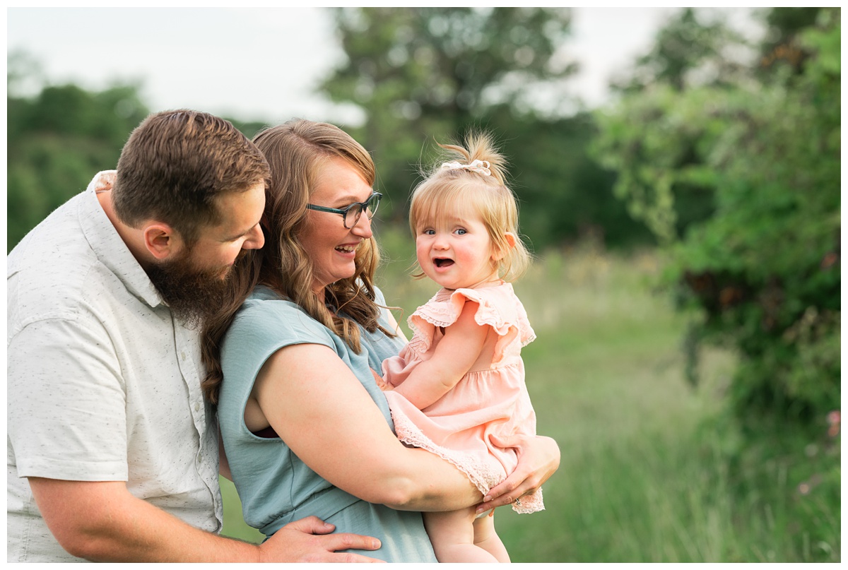 Family Photographers Columbus man in grey shirt hugs woman in blue dress holding baby girl in pink dress in green field