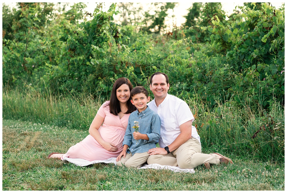 best maternity photographer new family of four sits in field with son holding wildflowers