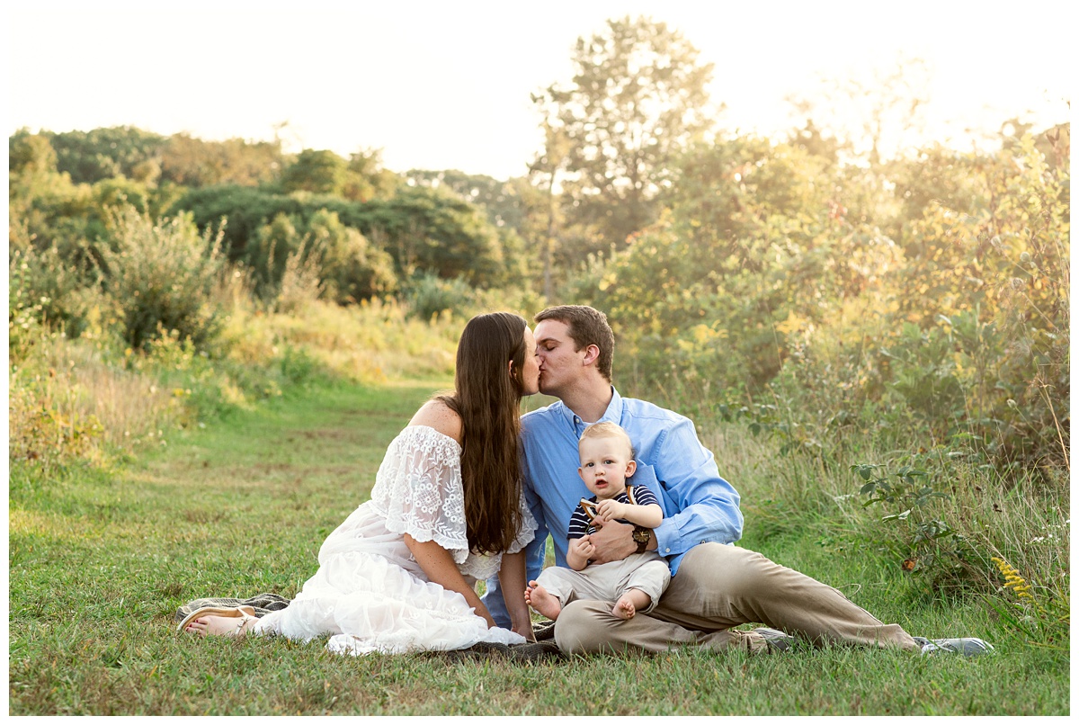 mom and dad kiss in field while baby boy looks on in Columbus Ohio